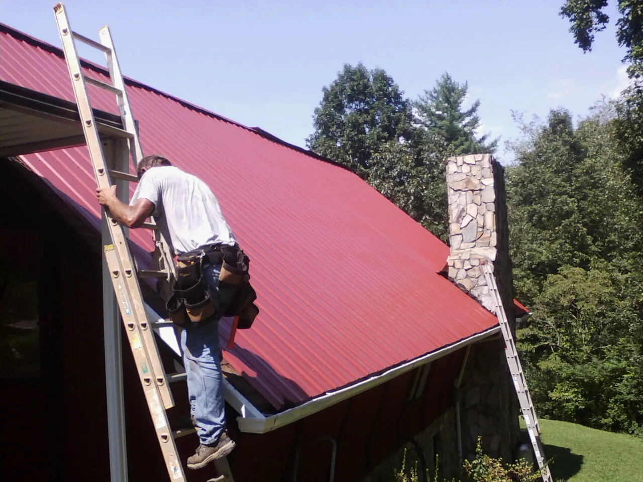 Roofer on a ladder beside a red metal roof and stone chimney — Soffit & Fascia Repair by Roofing Brothers in Carlstadt